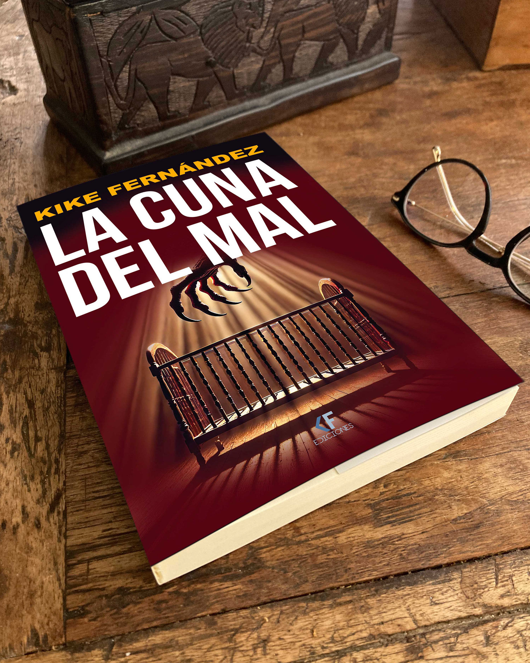 Portada de la novela La cuna del mal, de Kike Fernández, con una garra siniestra sobre una cuna iluminada, junto a unas gafas sobre una mesa de madera.