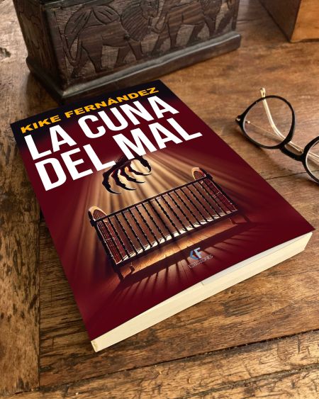Portada de la novela La cuna del mal, de Kike Fernández, con una garra siniestra sobre una cuna iluminada, junto a unas gafas sobre una mesa de madera.
